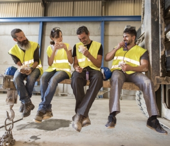 Vier Menschen sitzen in Arbeitskleidung auf einem Holzbalken und essen.