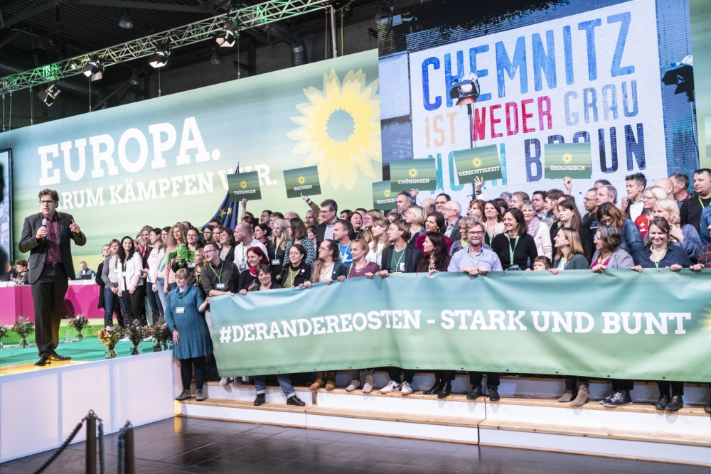 Eine Gruppe von grünen Parteimitgliedern stehen auf einer Treppe und halten auf der Bühne ein Banner vor sich. Banner-Text: "#DerAndereOsten - Stark und bunt."