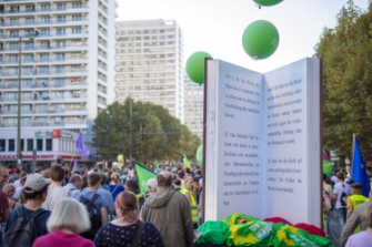 Eine demomenge zieht durch die Straßen. Die Menge ist von hinten fotografiert. Außerdem ist ein aufgeschlagenes Buch in Übergröße zu erkennen.