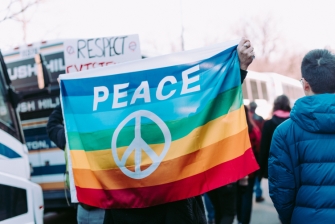Eine Person hält eine Flagge in Regenbogenfarben mit Peace-Zeichen und Aufschrift in die Kamera.