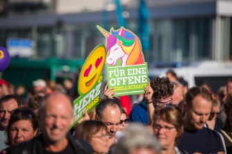Demonstrierende halten zwei Protestschilder in die Höhe. Schild 1: Smiley mit Herz-Augen, Text: "Herz statt Hass" / Schild 2: Buntes Einhorn, Text: "Für eine offene Gesellschaft"