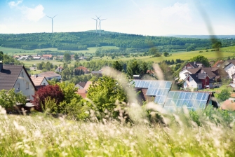 Eine Stadt in grüner Landschaft. Einige Häuser haben Solarzellen auf dem Dach, im Hintergrund sind Windenergieanlagen zu sehen.